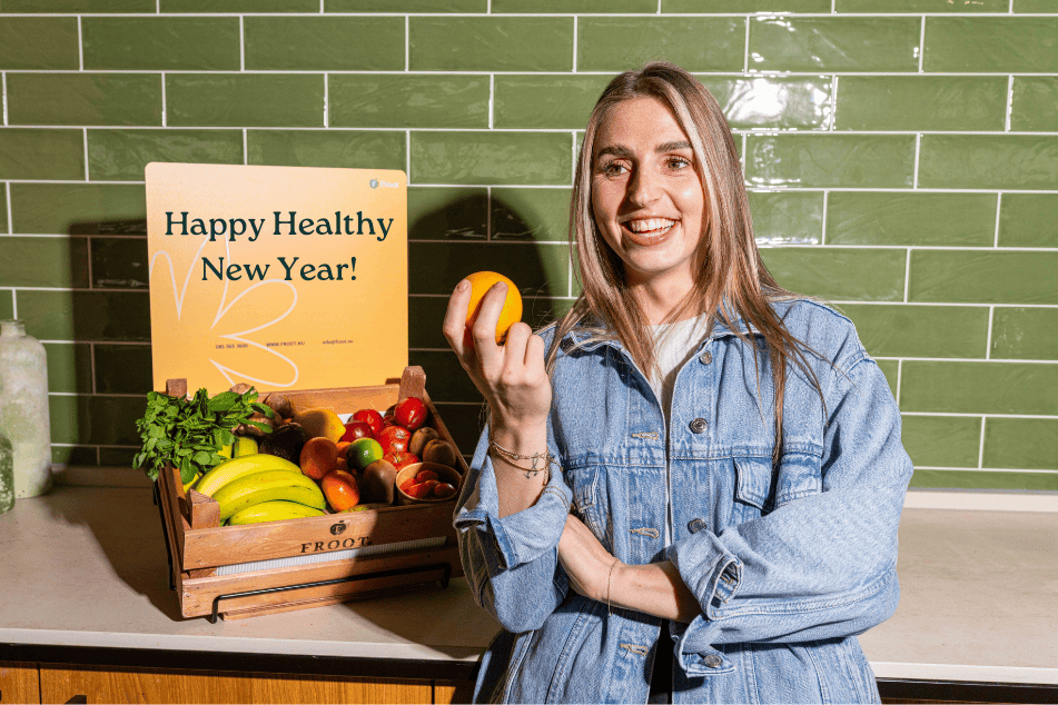 Vrolijke medewerker met een stuk fruit in de hand naast een Froot-Krat vol vers fruit op kantoor, gebruikt voor vitaliteit en gezonde voornemens in 2026.