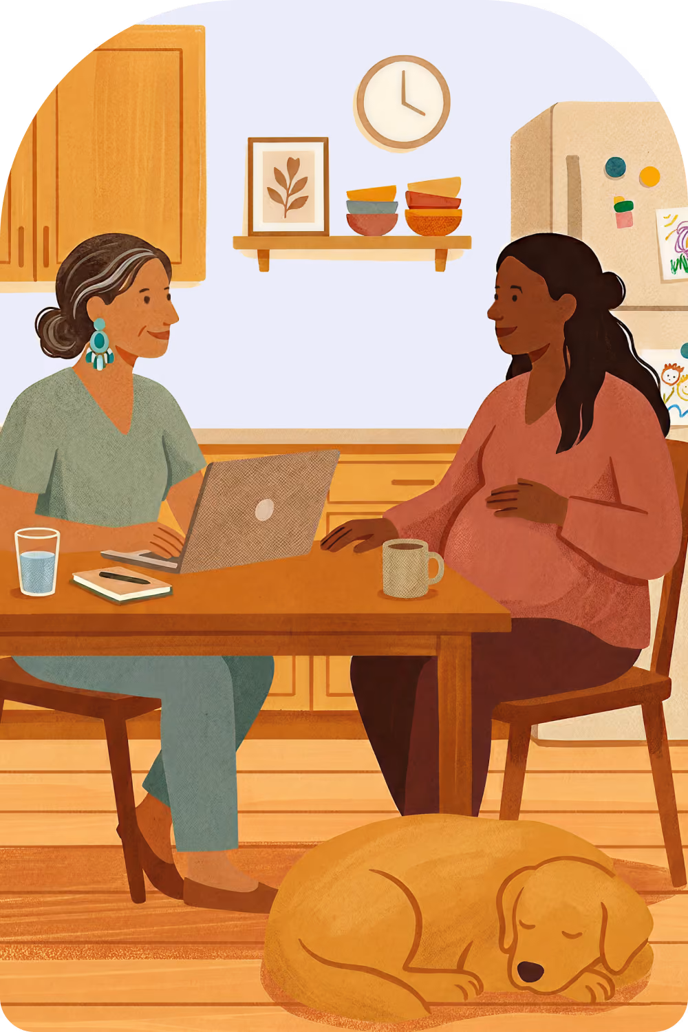 Two women sitting at a kitchen table with a laptop and mugs while a dog sleeps on the floor.