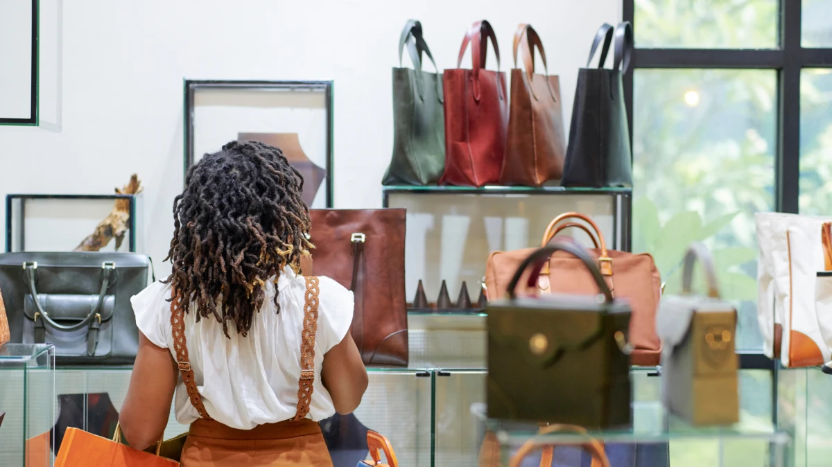 Pensive stylish young woman looking at various leather bags on shelves in department store