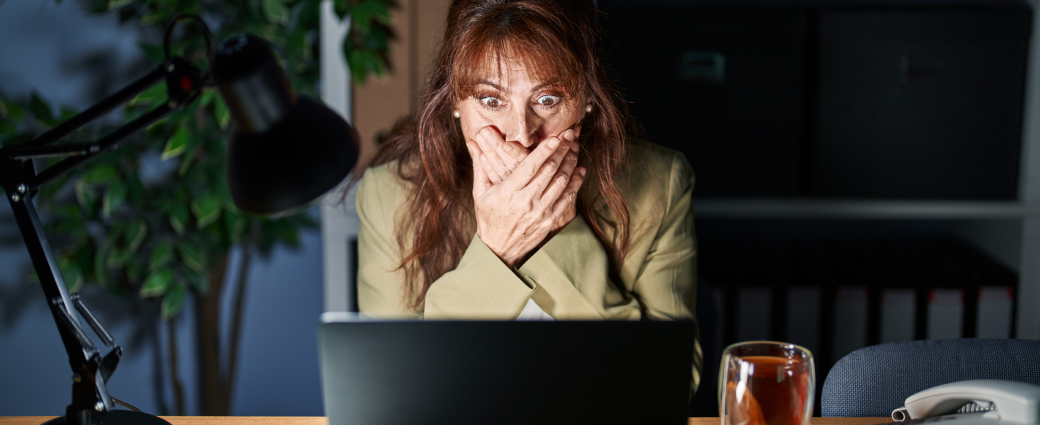 A woman covers her mouth with her hands as she gasps at her laptop screen.