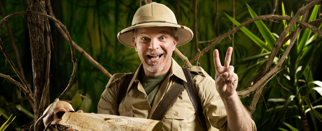 A smiling man in safari gear and holding a treasure map gives the viewer a peace sign.