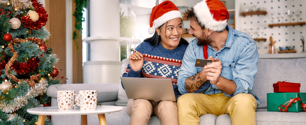 A woman and man wearing Santa hats huddle around a laptop, waving a credit card and shopping