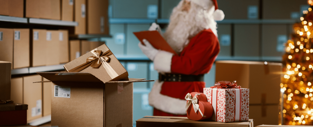 Packages sit next to a Christmas tree in a store room while Santa checks inventory on a clipboard in the background.