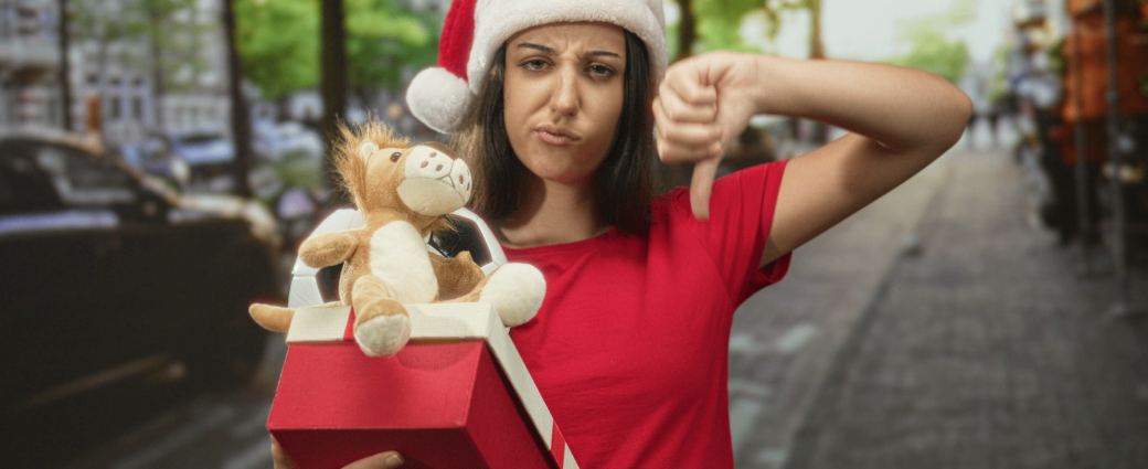A woman with a disappointed face and wearing a Santa hat holds an open Christmas gift and shows a thumbs down.