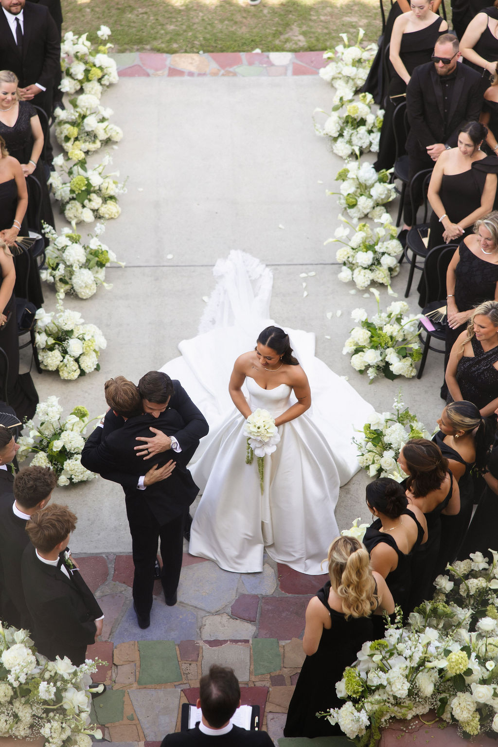 Overhead view of a Florida wedding ceremony with a bride holding a white bouquet and an aisle lined with lush white floral arrangements by ImpeKable Flowers & Event Design.