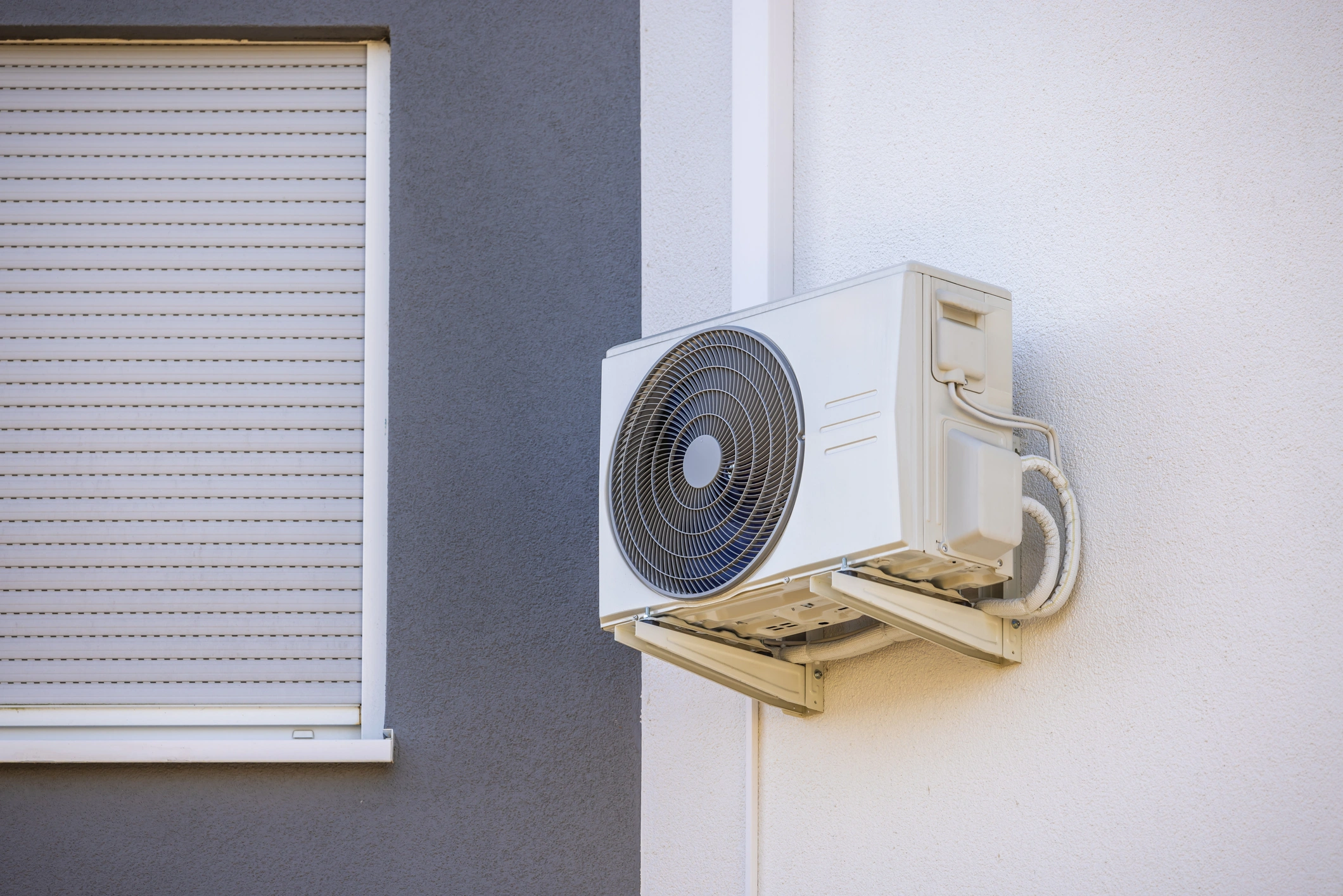 Wall-mounted air conditioner on a white facade, adjacent to a window with a closed gray shutter. 