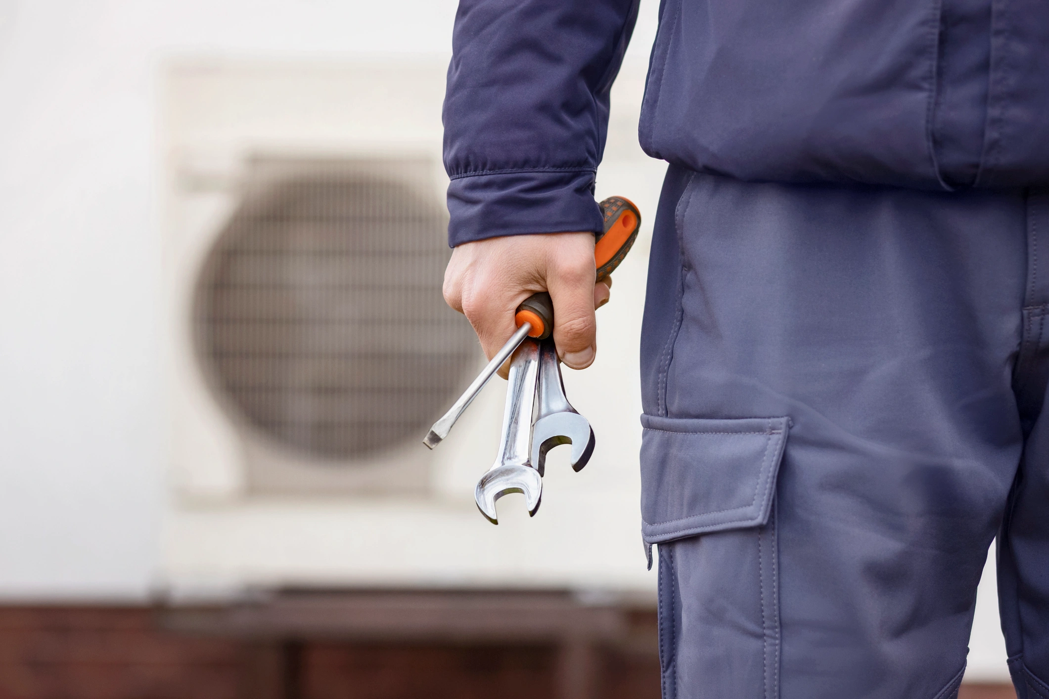 A worker in a blue uniform holds wrenches and a screwdriver. In the background, an air conditioning unit is visible.