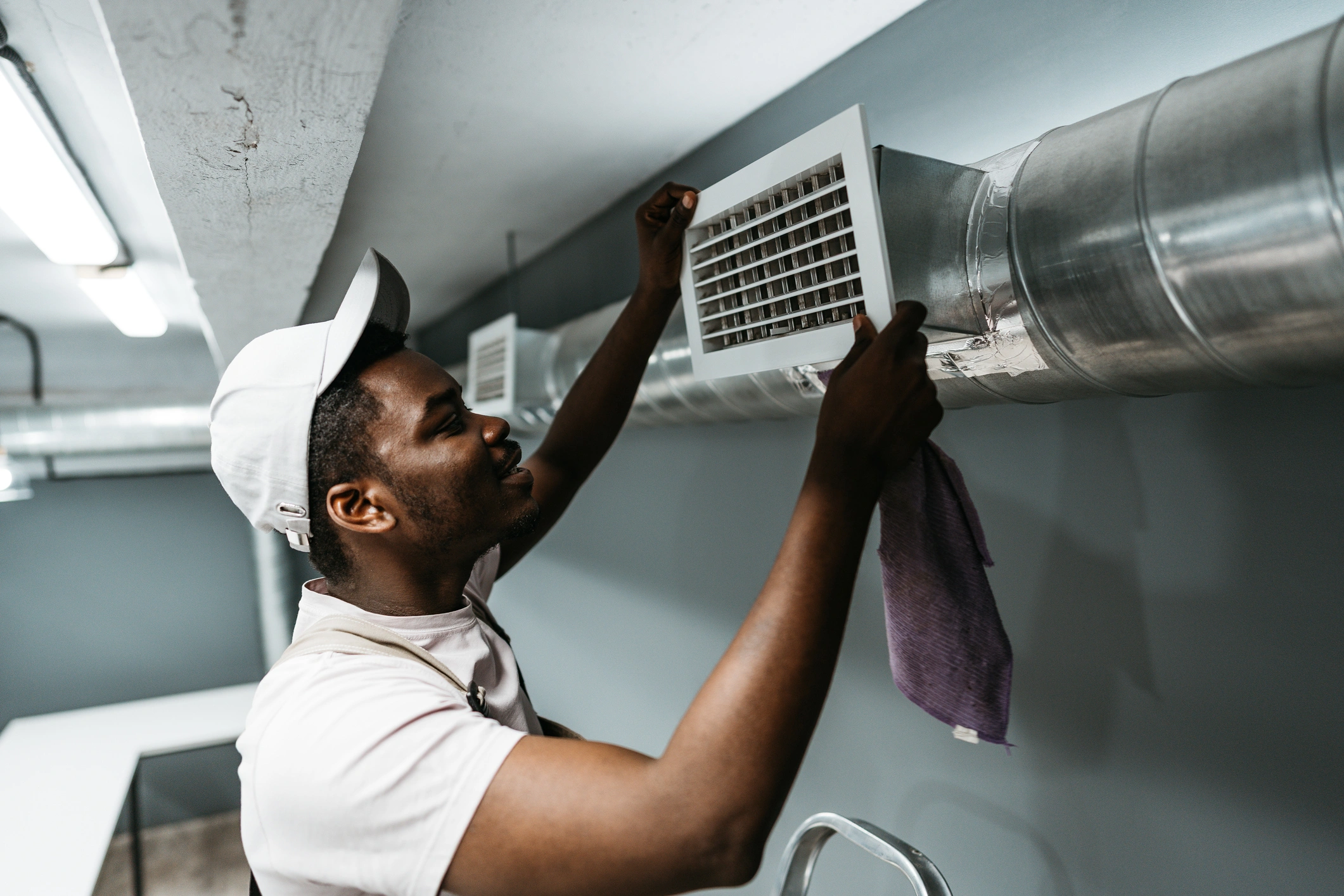A man in a white shirt and cap adjusts a vent on a metal air duct in a modern room. 