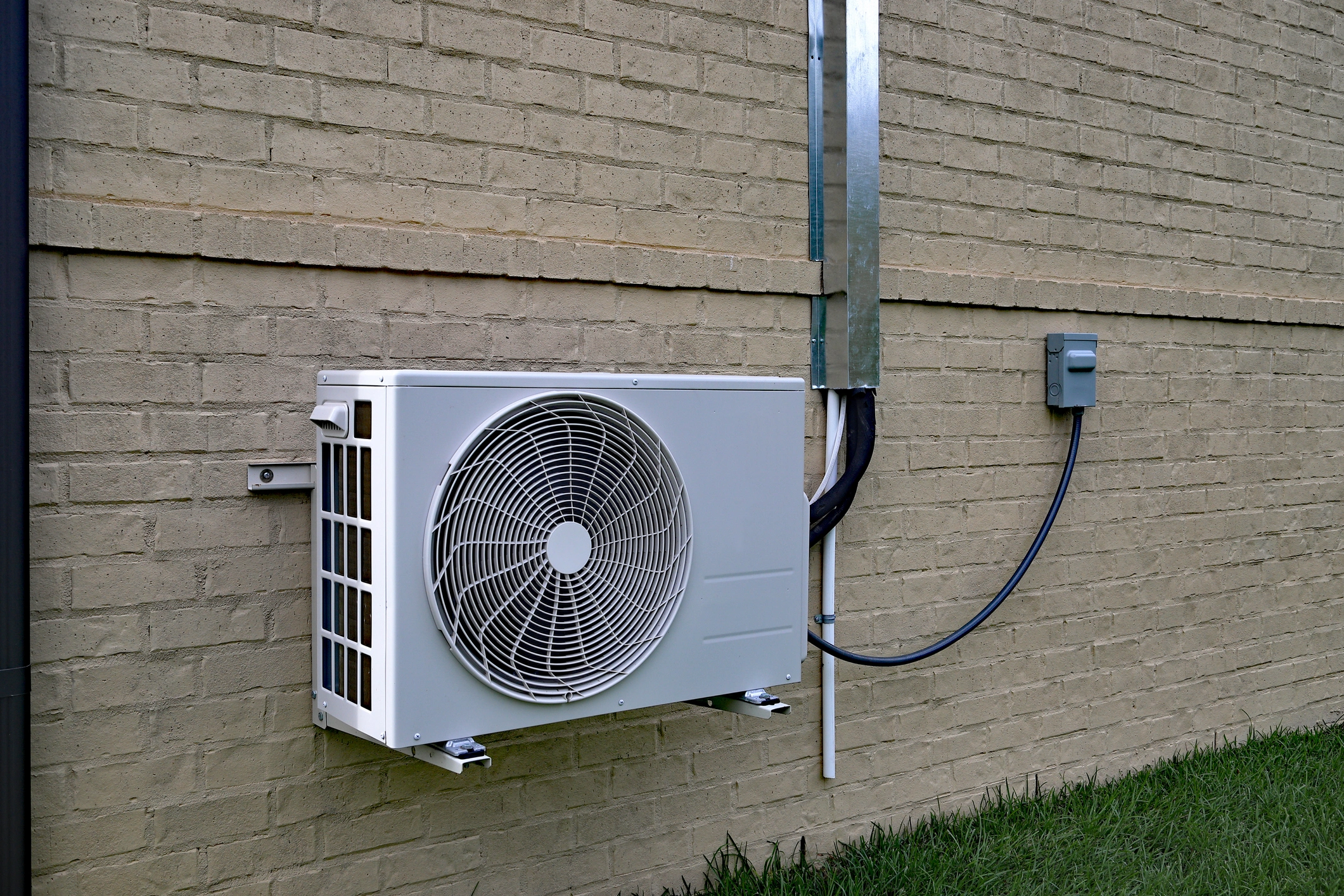 Outdoor air conditioning unit mounted on a beige brick wall with visible electrical cables and conduit