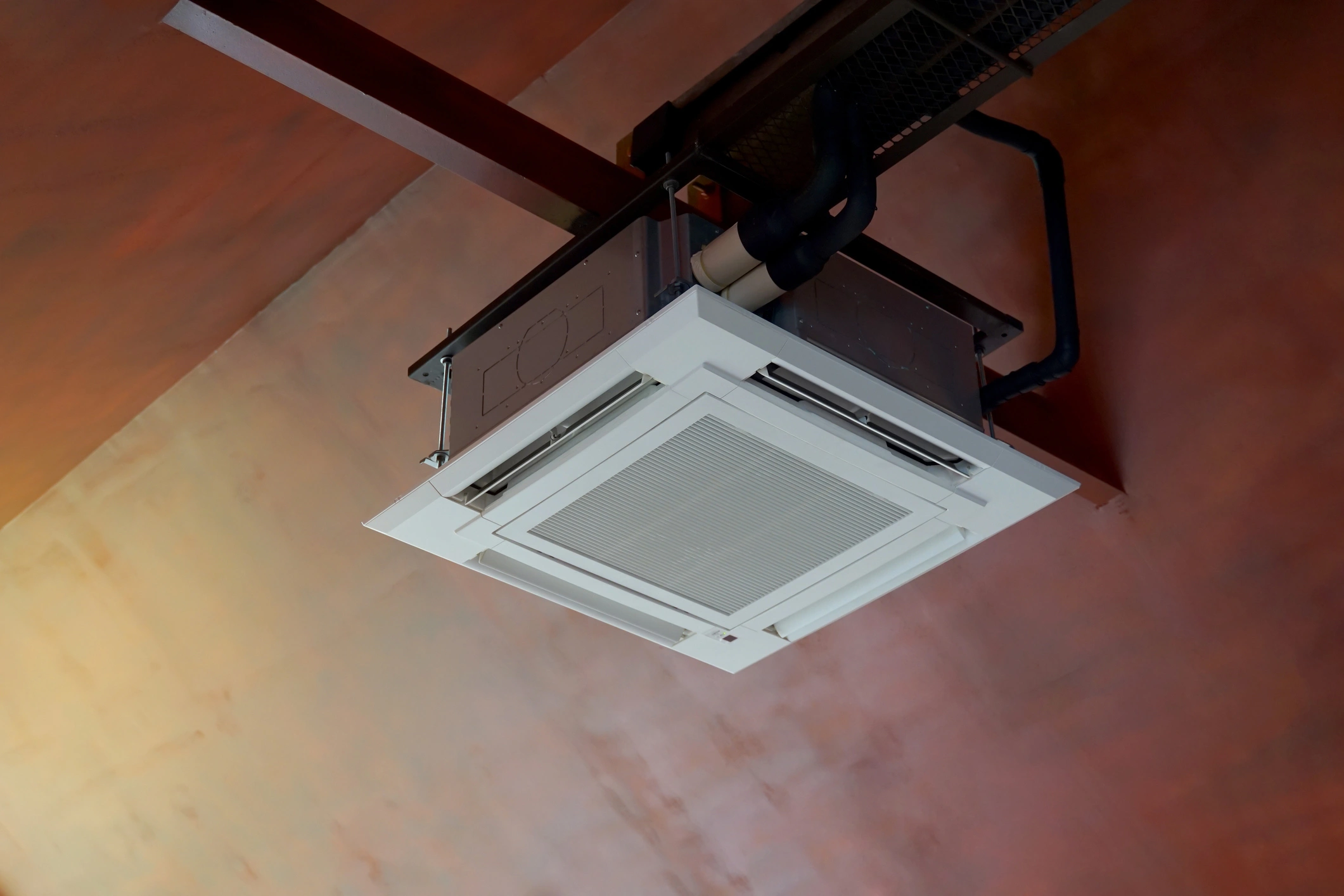 Ceiling-mounted air conditioning unit with visible ducts on a reddish-brown ceiling. 