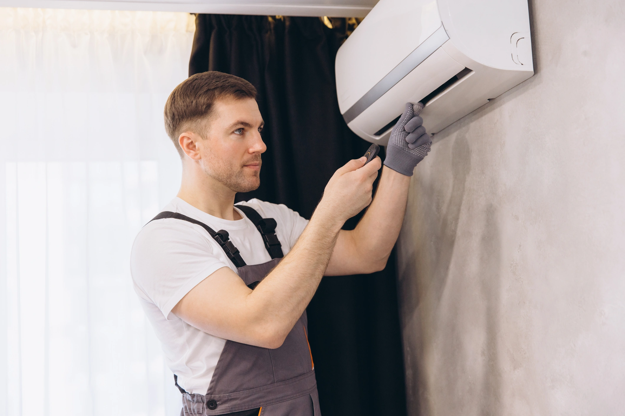A technician wearing gloves and overalls adjusts a wall-mounted air conditioner. 