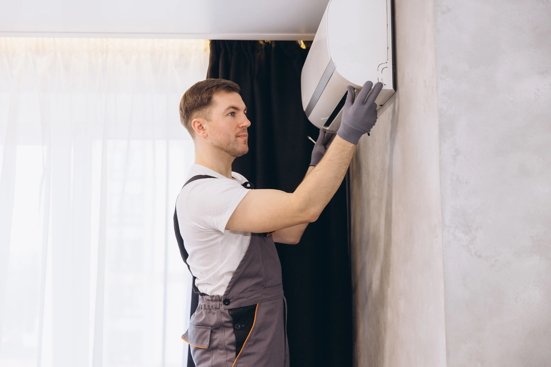 A technician in overalls and gray gloves adjusts a wall-mounted air conditioner. 