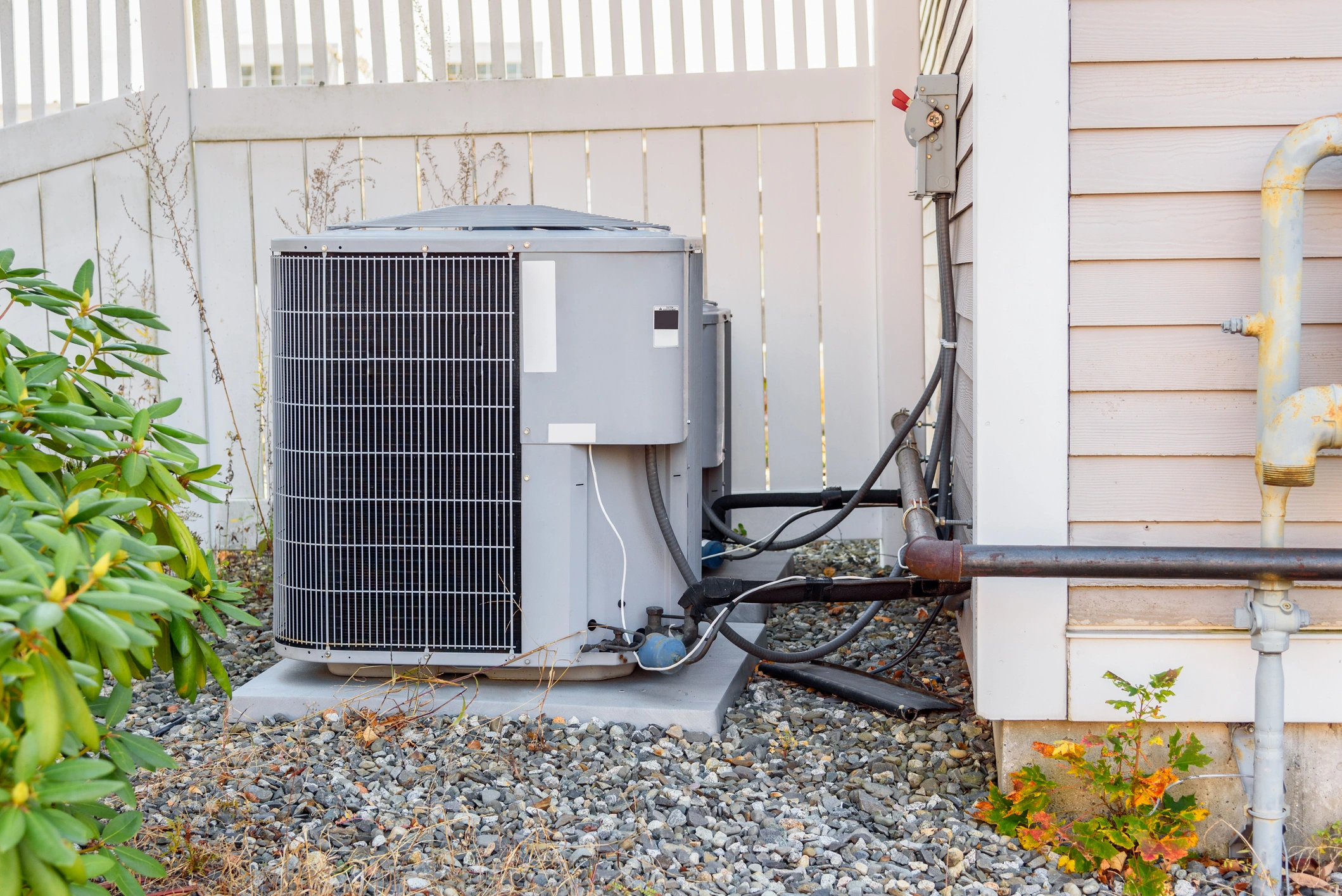 Outdoor air conditioning unit next to a house with siding, surrounded by a white fence.