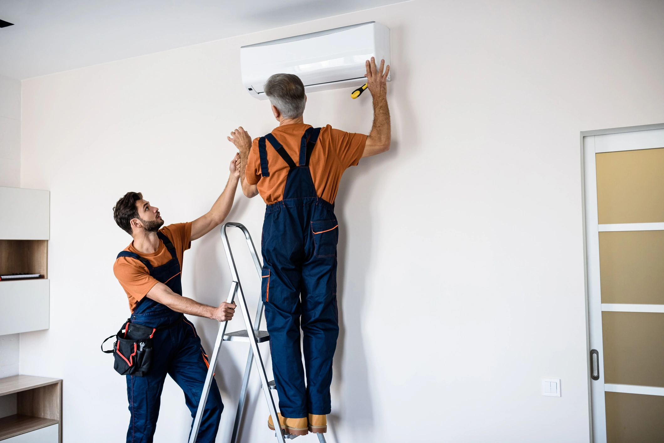 Two technicians in orange shirts and blue overalls install an air conditioner. 
