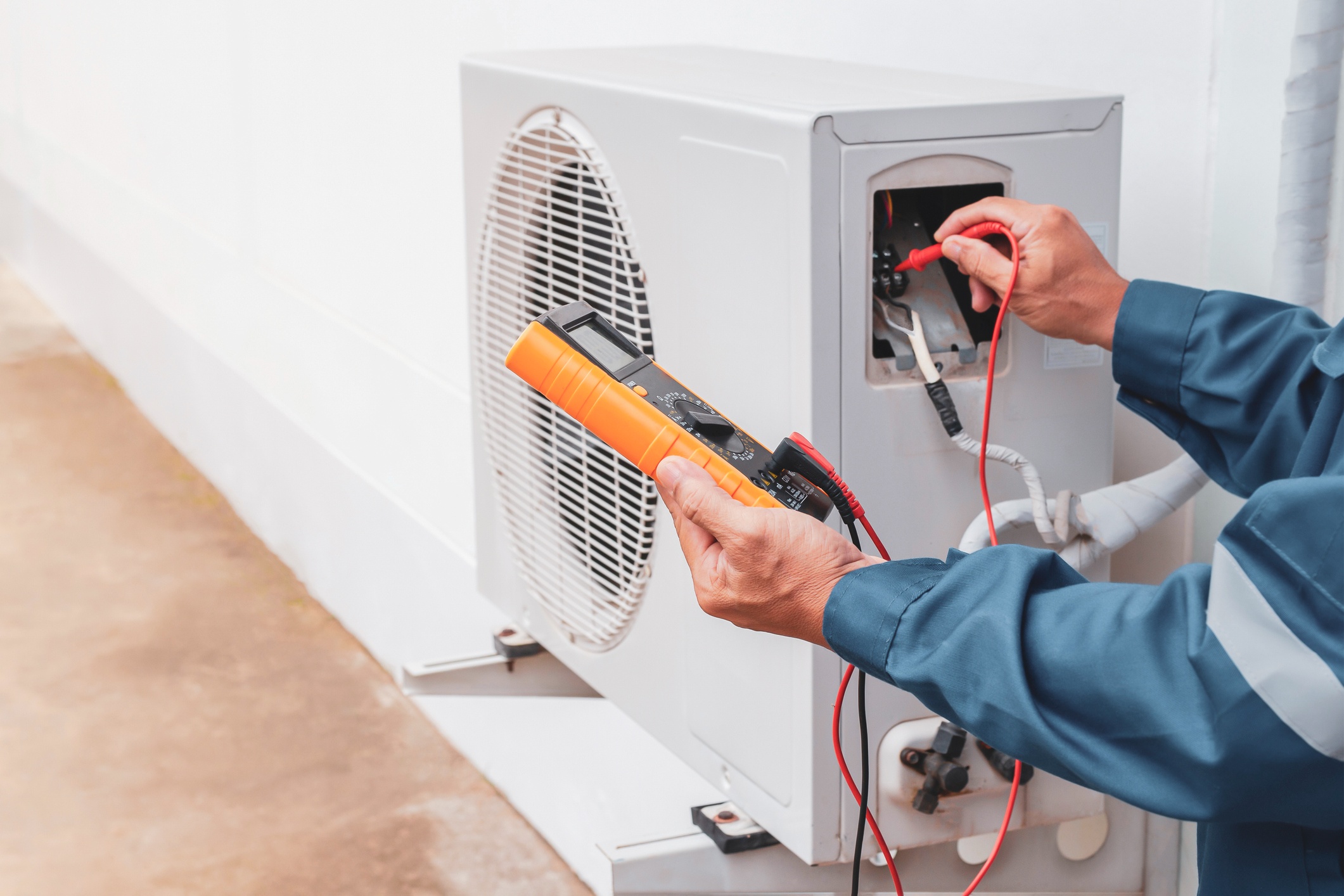 A person in blue work attire is using a multimeter to check the electrical connections on a white outdoor air conditioning unit.