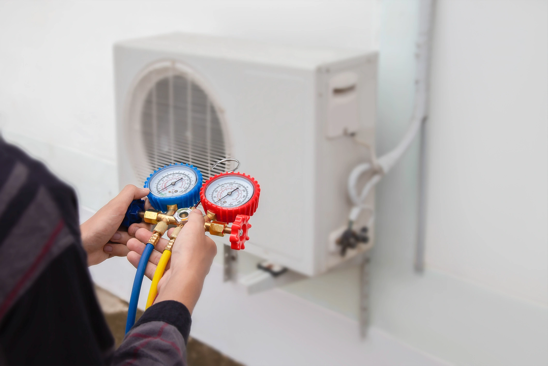A person holds red and blue manifold gauges in front of an outdoor air conditioning unit