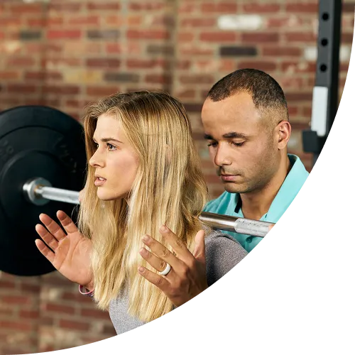 A physical therapist in a teal polo shirt closely observes and guides a patient performing a barbell squat in a gym setting.