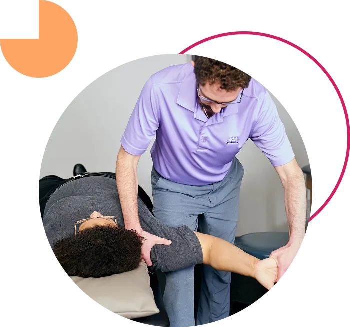 A physical therapist in a light purple polo shirt provides hands-on treatment, assisting a patient with a shoulder and arm mobility exercise on a treatment table.