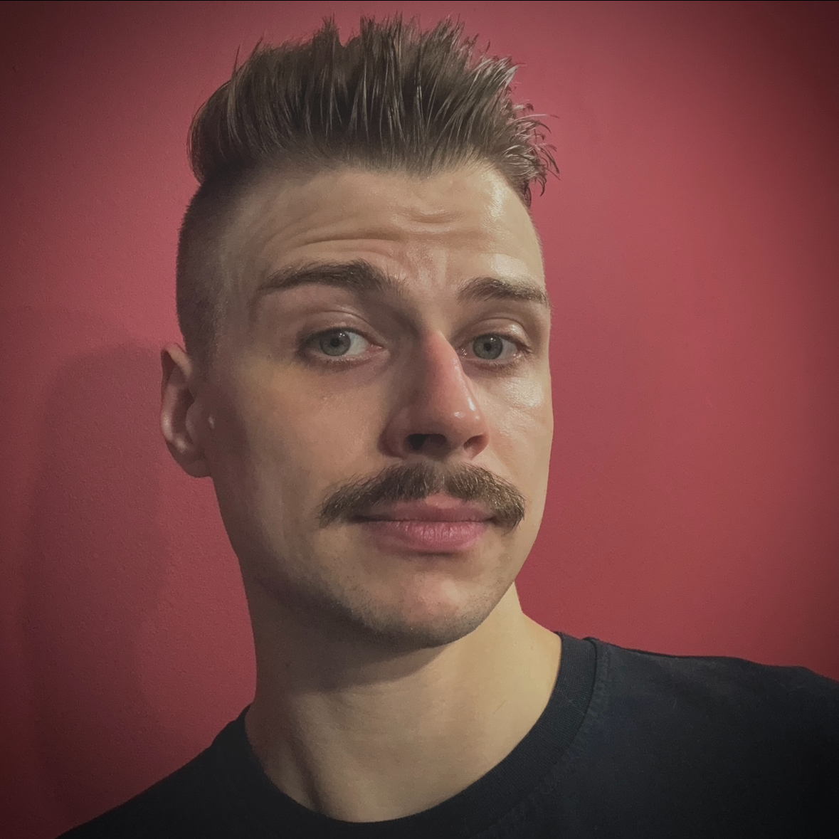 Young man with a styled short haircut and a mustache posing against a red background.