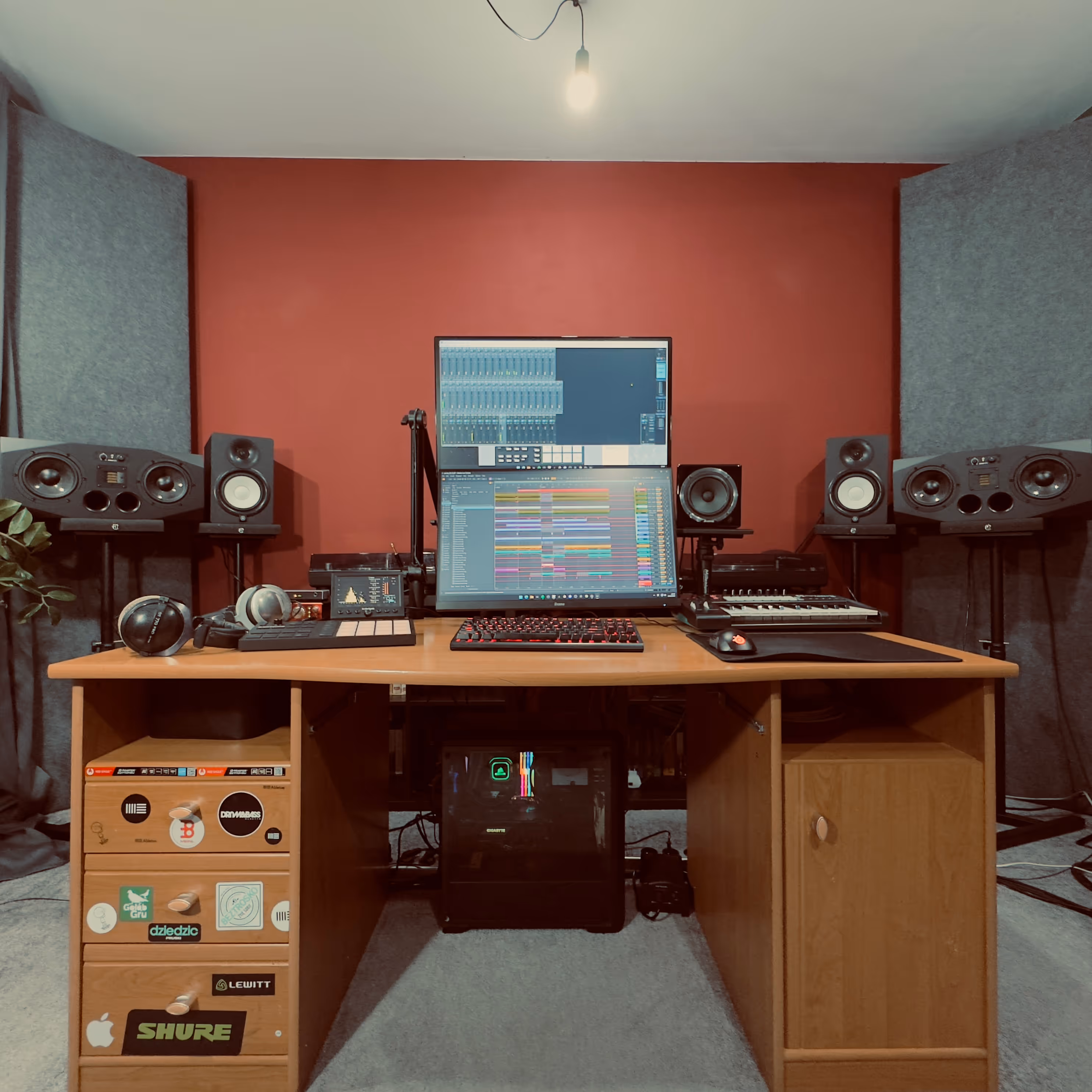 Home music production setup with dual monitors, keyboard, headphones, speakers, and soundproofing panels on a red wall.