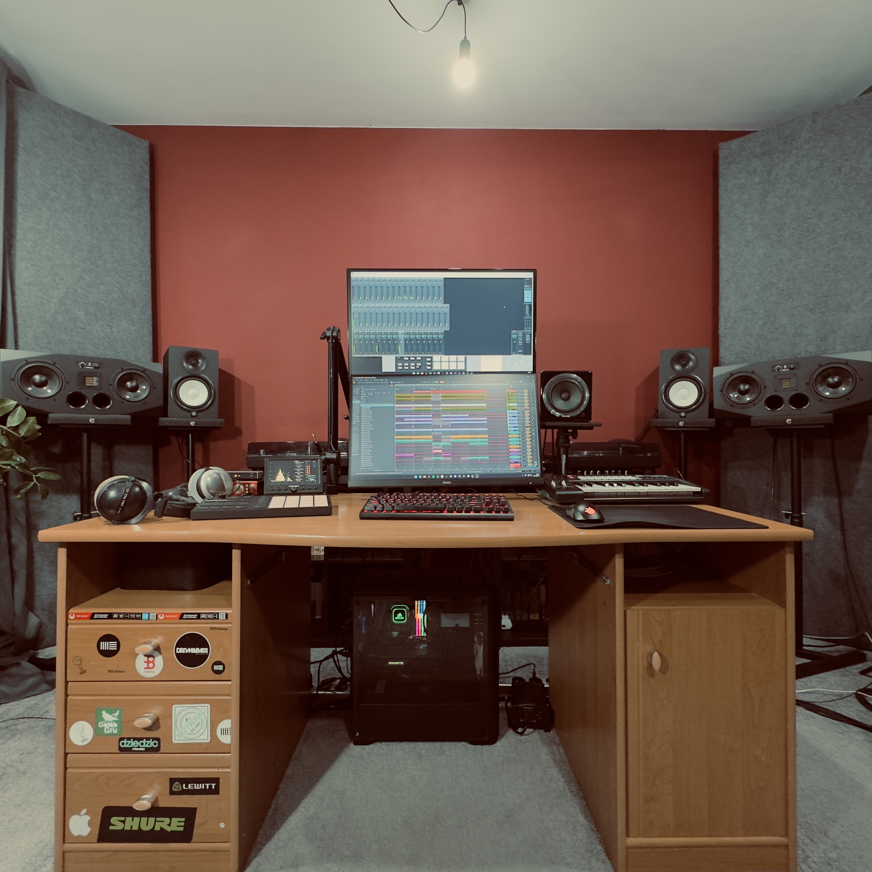 Home music production setup with dual monitors, keyboard, headphones, speakers, and soundproofing panels on a red wall.