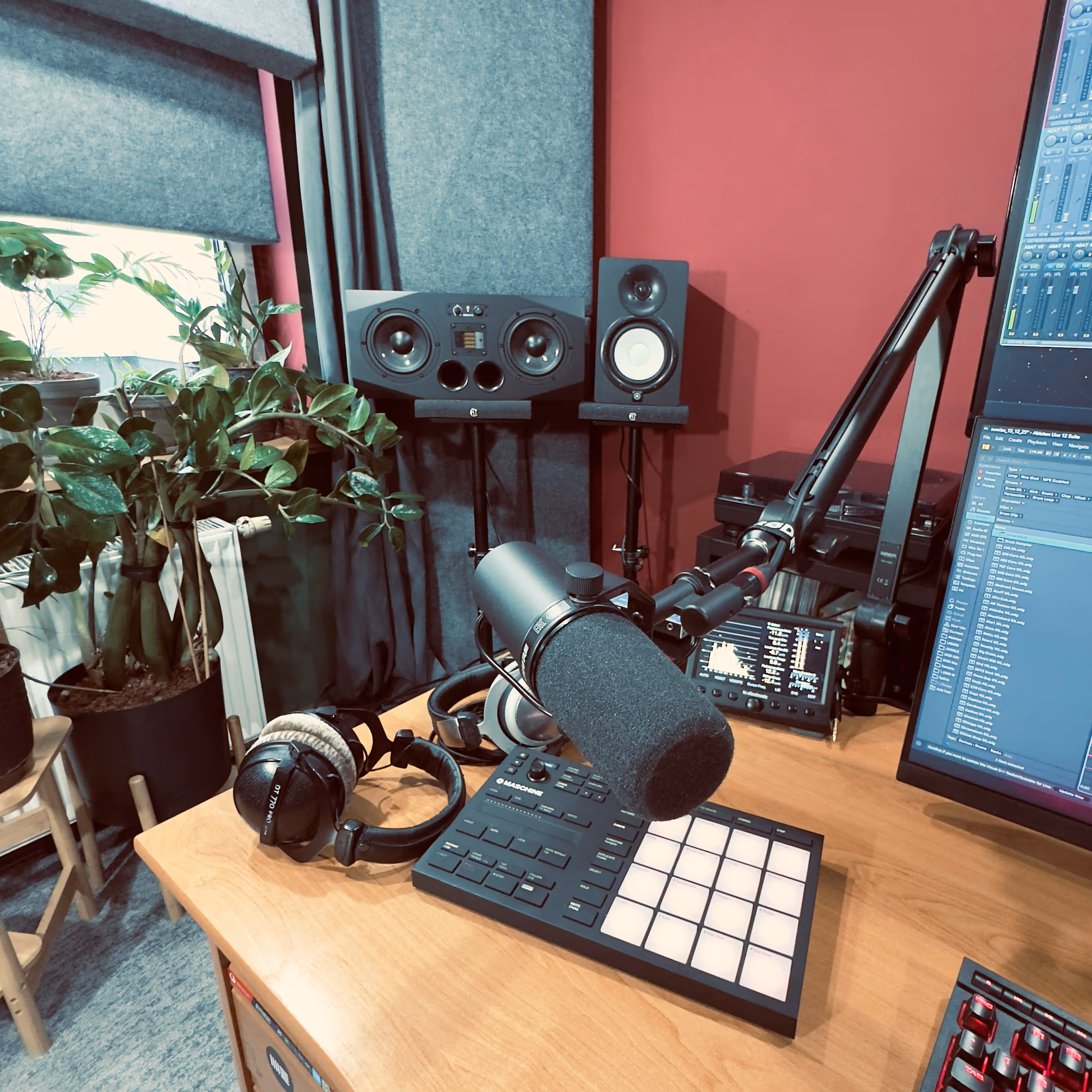 Podcast or music production desk with a professional microphone, headphones, music sampler, studio monitors, and computer screens in a room with plants and red walls.