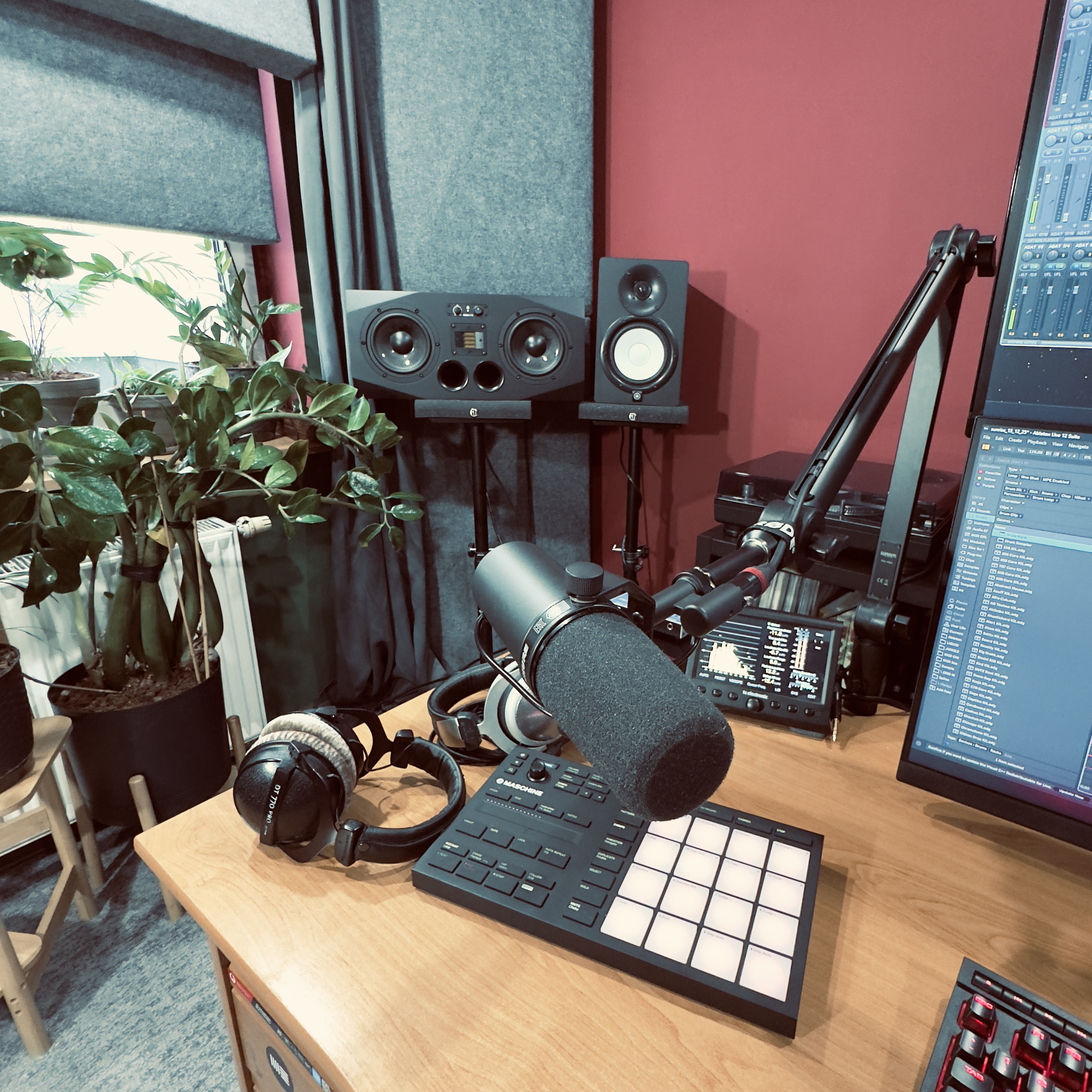 Podcast or music production desk with a professional microphone, headphones, music sampler, studio monitors, and computer screens in a room with plants and red walls.
