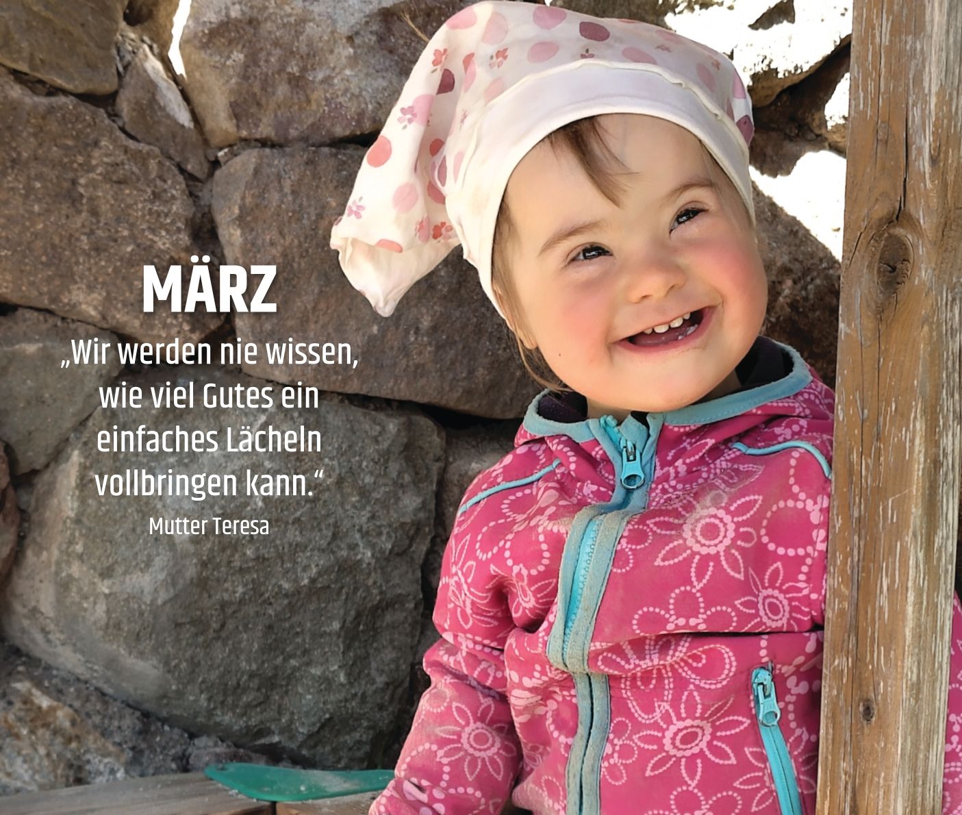 Kleines Mädchen mit Trisomie 21 sitzt auf einer Bank vor einer Mauer und lacht fröhlich in die Kamera.