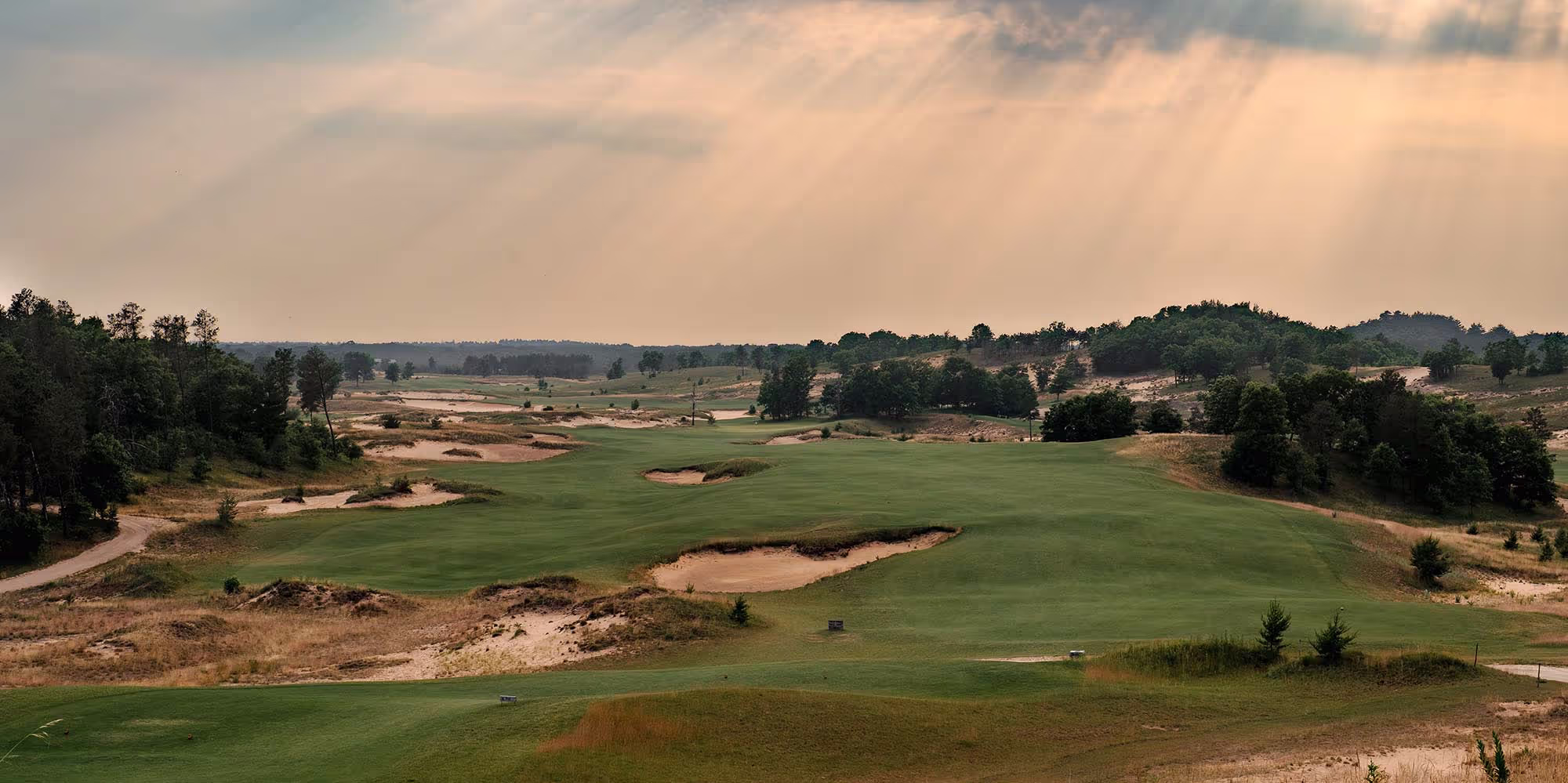 Wide view of a golf course at sunrise or sunset, with rays of light streaming through clouds over rolling fairways, bunkers, and surrounding trees.