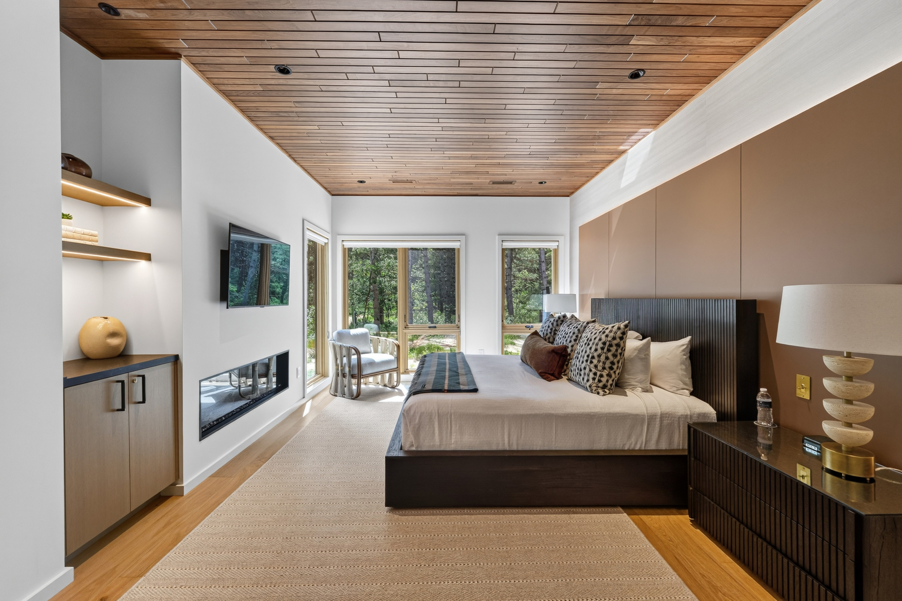 Bedroom with wood ceiling, large bed, fireplace, and forest views.