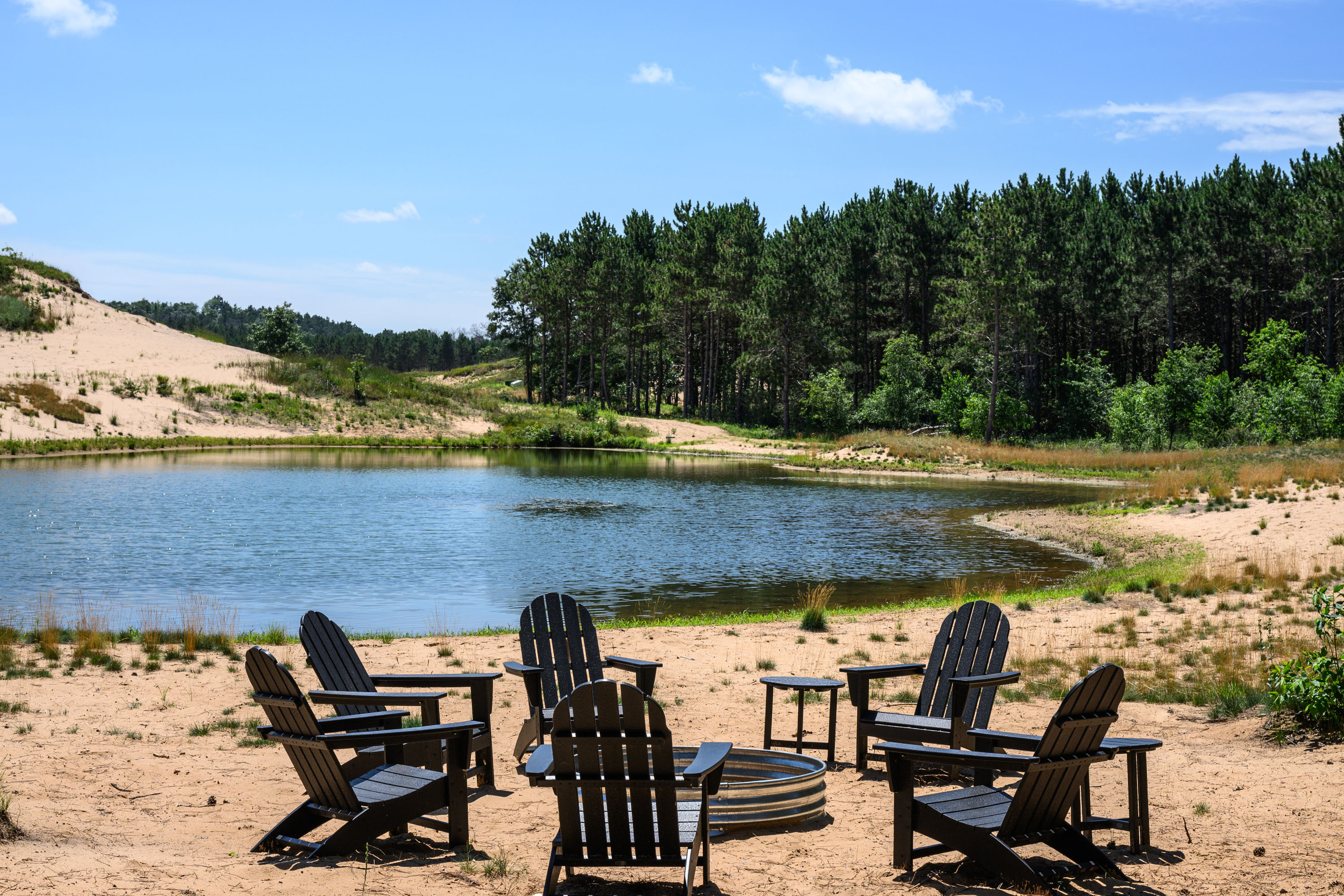 Adirondack chairs surrounding a fire pit beside a pond and sandy dunes.