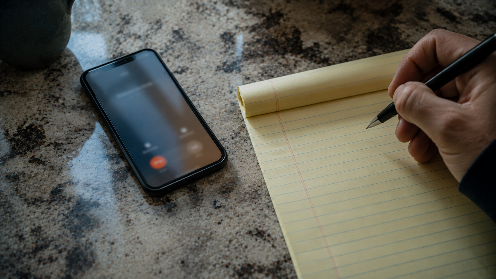 A phone mid call on a kitchen counter beside a legal pad, with a pen hovering above the page.