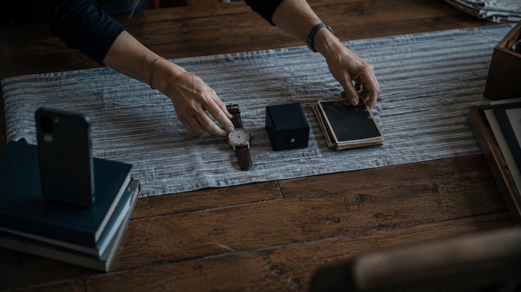 Hands arranging a watch, a jewelry box and face down photographs into a grid on a dining table.