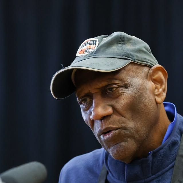 Spencer Haywood in a Basketball Hall of Fame cap speaking into a microphone at a press event.