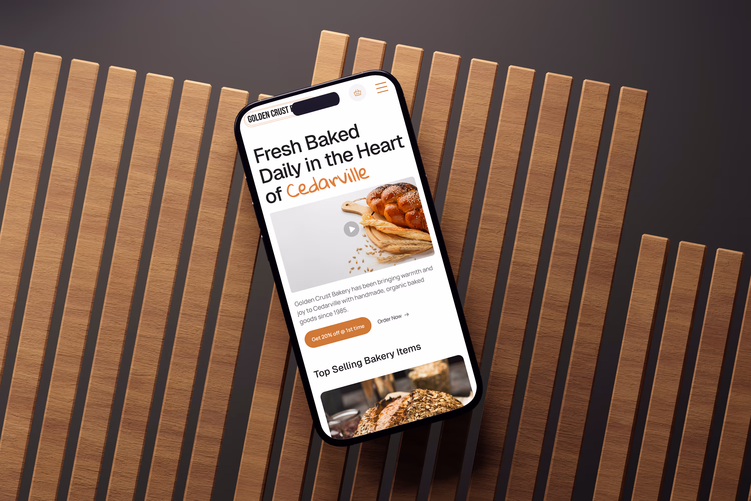 Smartphone displaying a bakery website with featured bread and Cedarville branding, placed on a dark wooden slat background for a warm, modern aesthetic.