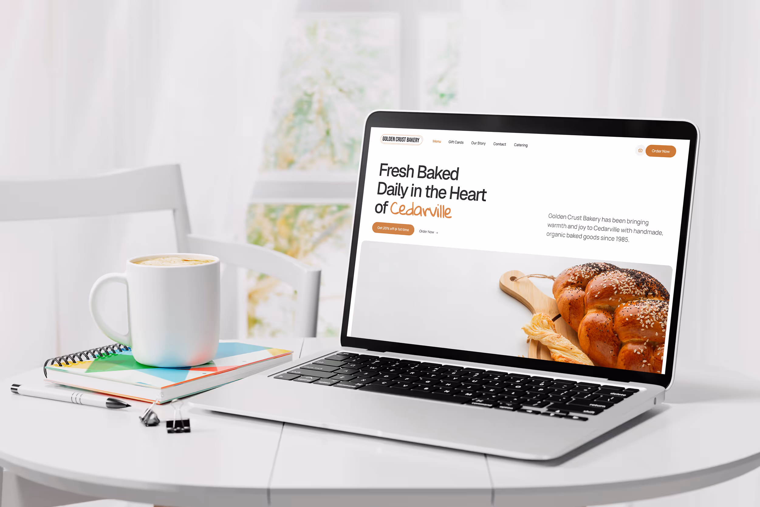 Laptop on a white table displaying a bakery homepage with organic bread, alongside a coffee mug and colorful notepad in a bright, cozy setting.