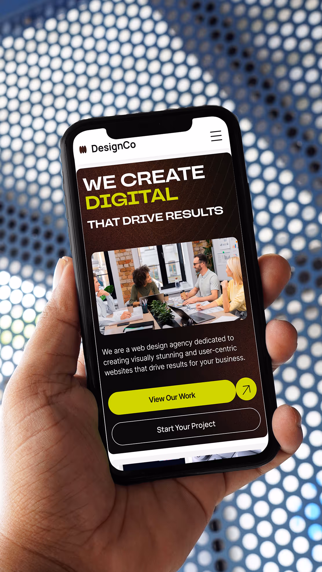 Hand holding a smartphone displaying the DesignCo web agency homepage with bold headline and yellow CTA, against a blue industrial mesh background.