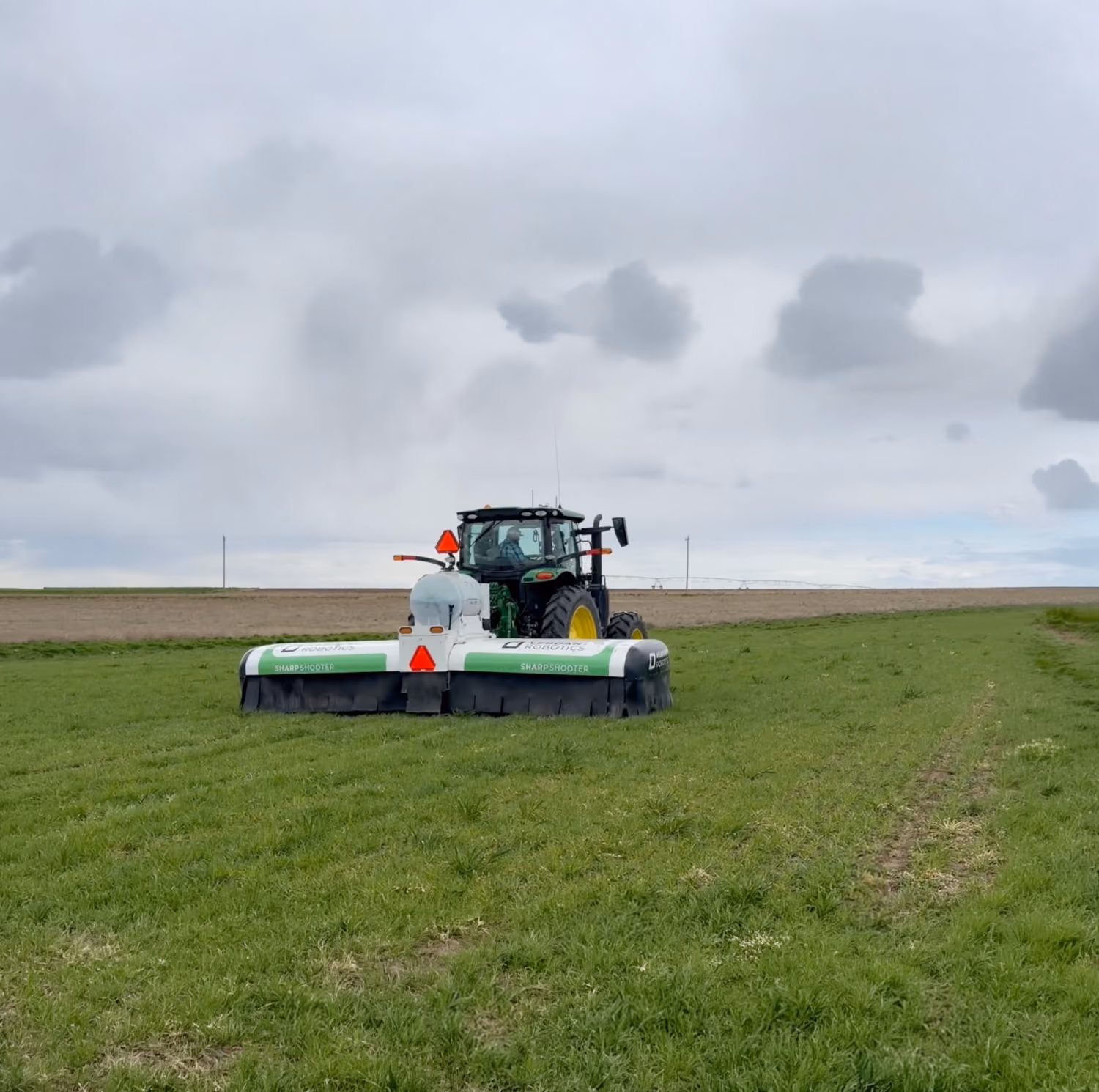 Tractor with attached SharpShooter sprayer working on a green agricultural field under cloudy sky.