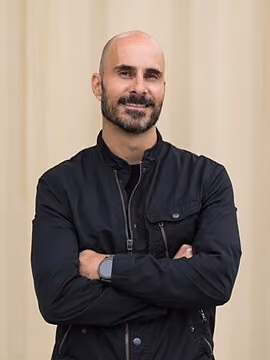 Man with a bald head and beard smiling with arms crossed, wearing a black jacket, standing against a beige background.