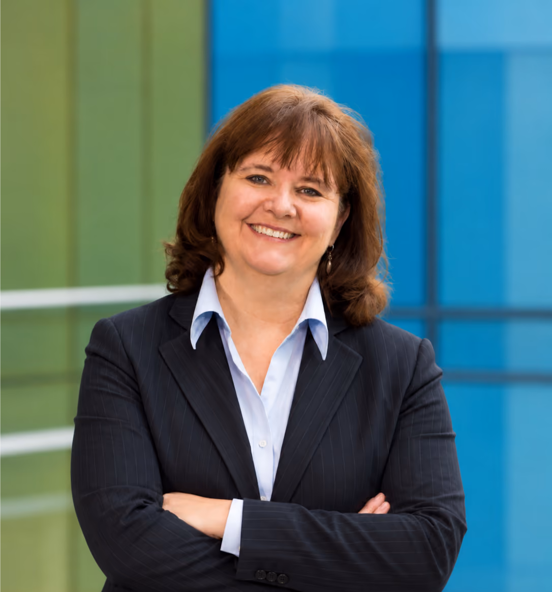 Smiling woman with medium-length brown hair wearing a dark pinstripe blazer and white collared shirt, standing against a blue and green background.
