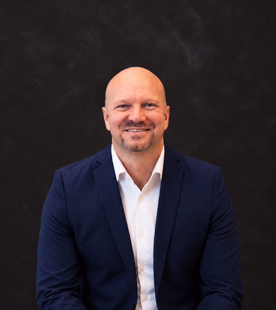 Smiling bald man with a trimmed beard wearing a white shirt and dark blazer against a dark background.