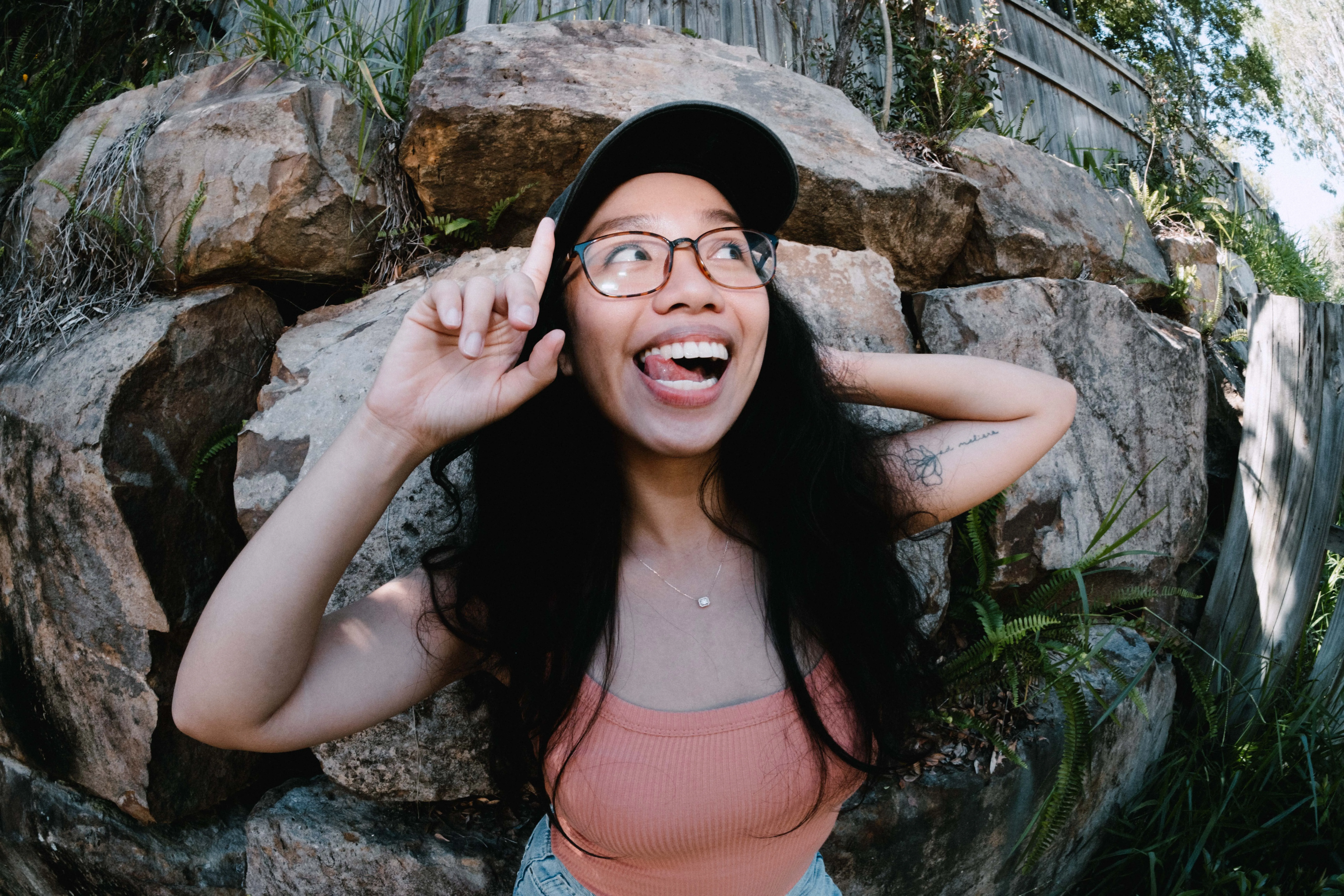 joyful young woman with long dark hair, glasses, and a cap playfully points upward while sticking out her tongue. She’s wearing a peach tank top and standing in front of large rocks and greenery, smiling with an expressive and carefree pose