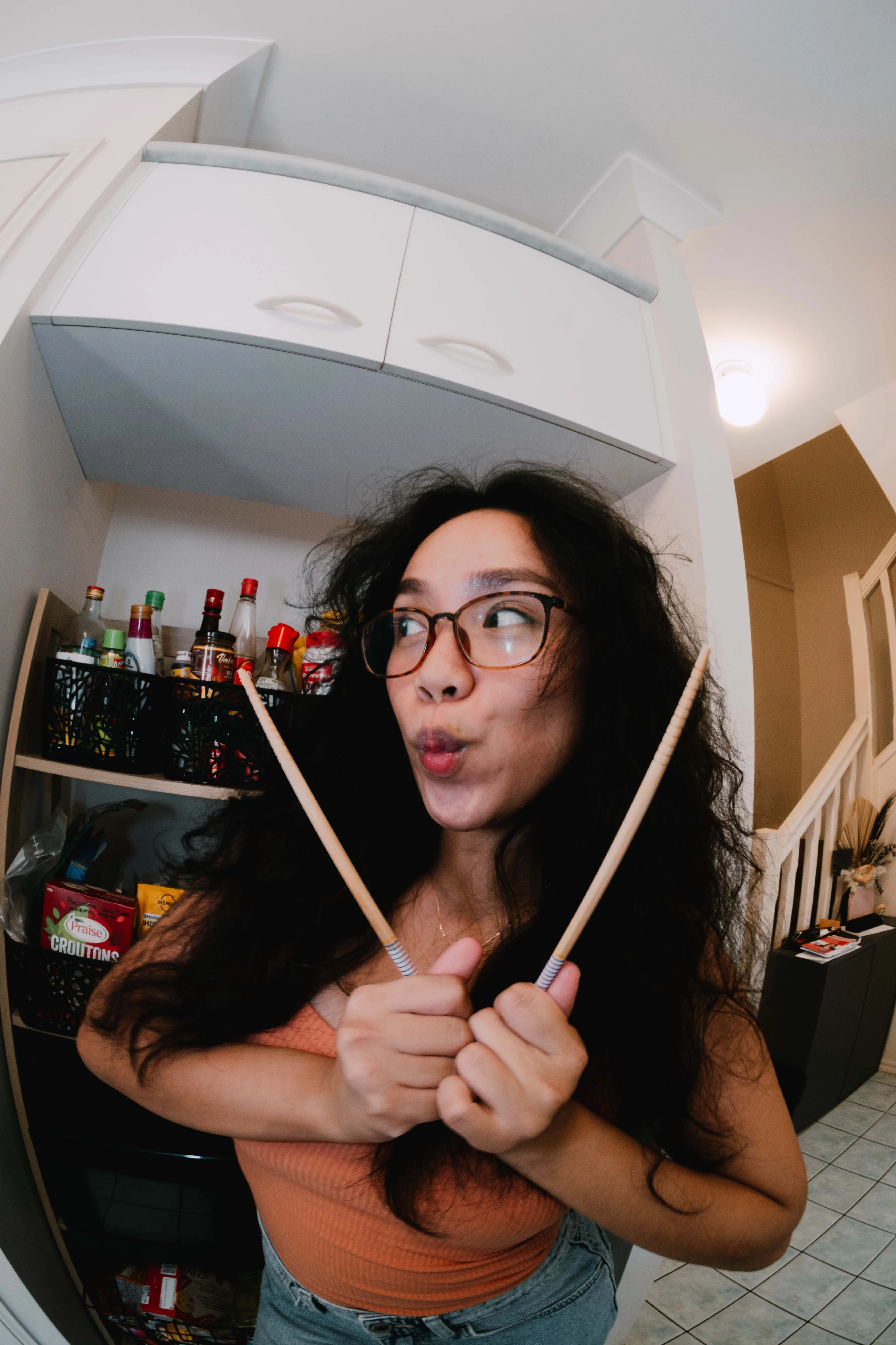 A woman wearing glasses and pursing her lips, holding up two chopsticks near her face, forming a V-shape