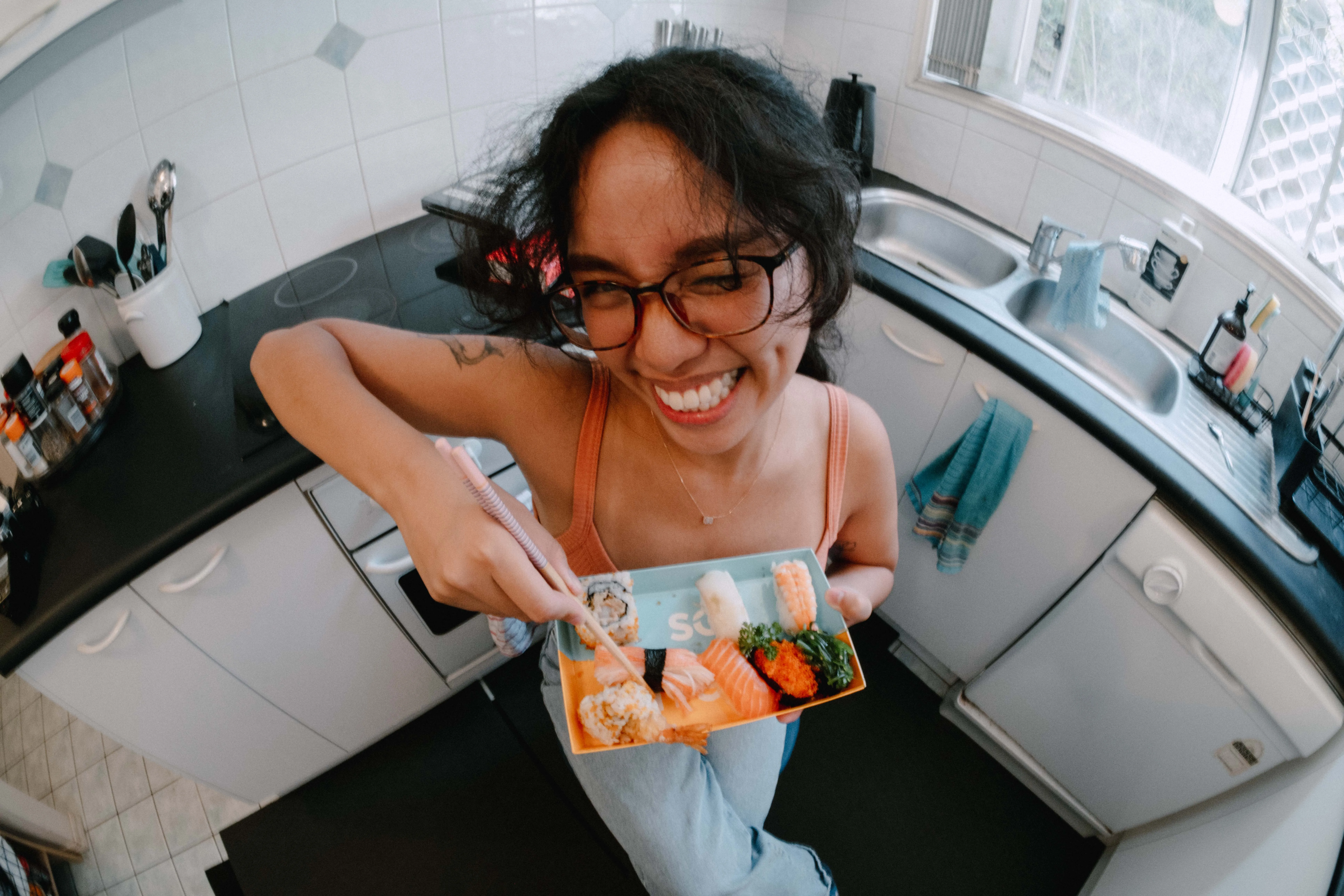 joyful young woman with long dark hair, glasses, and a cap playfully points upward while sticking out her tongue. She’s wearing a peach tank top and standing in front of large rocks and greenery, smiling with an expressive and carefree pose