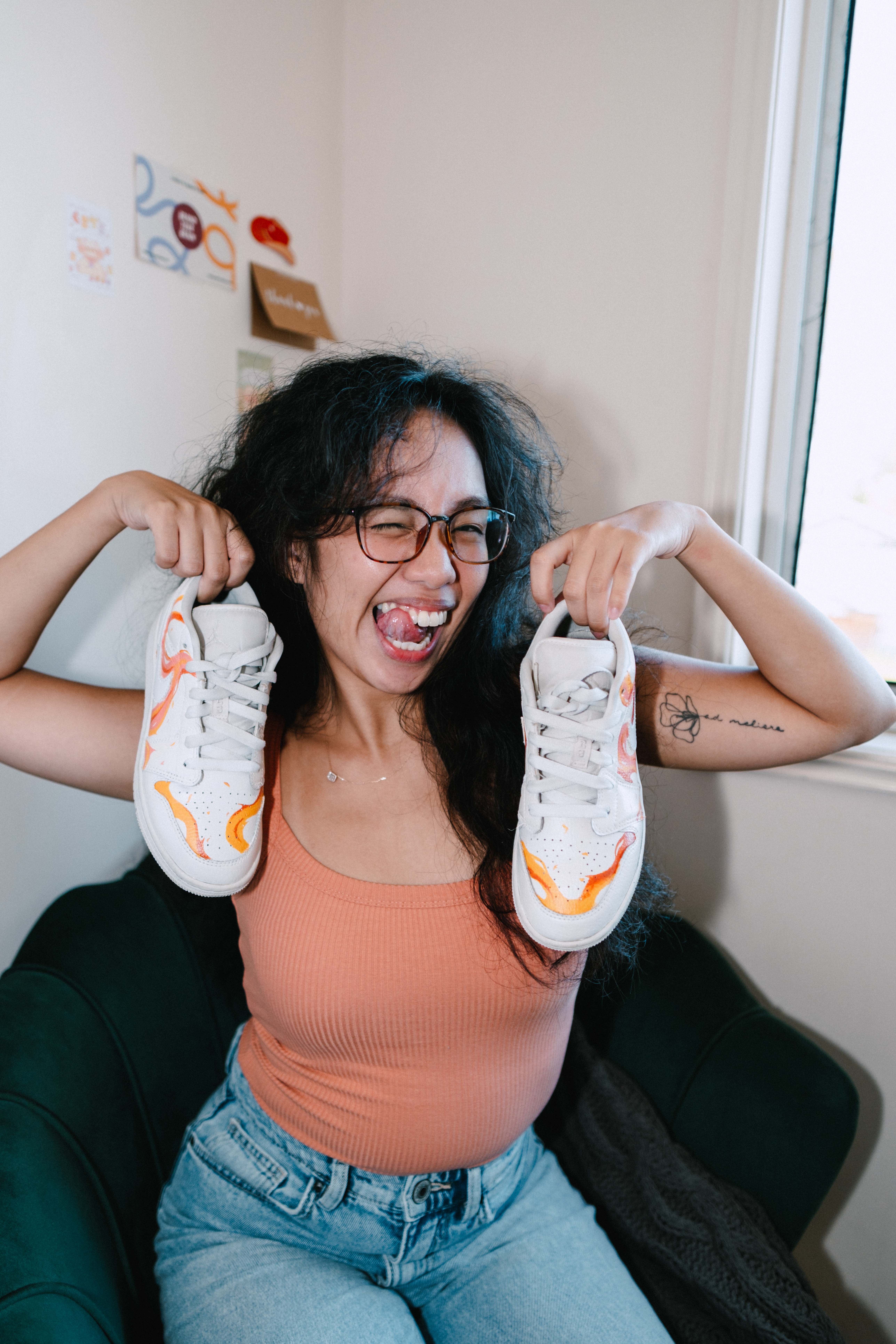 joyful young woman with long dark hair, glasses, and a cap playfully points upward while sticking out her tongue. She’s wearing a peach tank top and standing in front of large rocks and greenery, smiling with an expressive and carefree pose