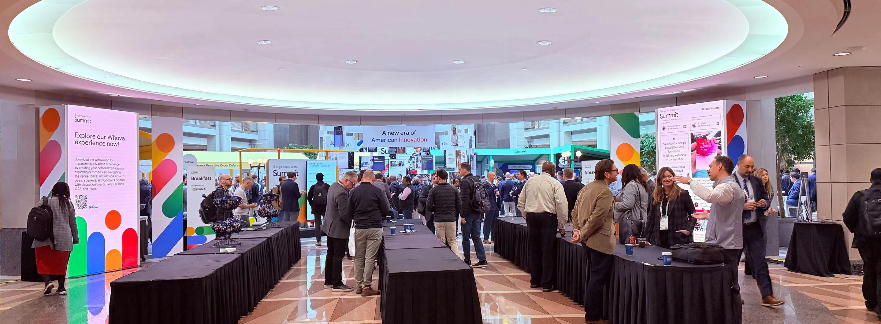 Crowd of people gathered at a conference or summit with colorful informational booths and banners in a spacious indoor venue.
