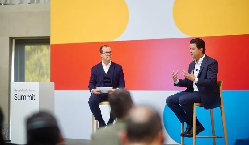Two men in suits seated on stools having a discussion on stage at the Google Public Sector Summit with a colorful abstract background.