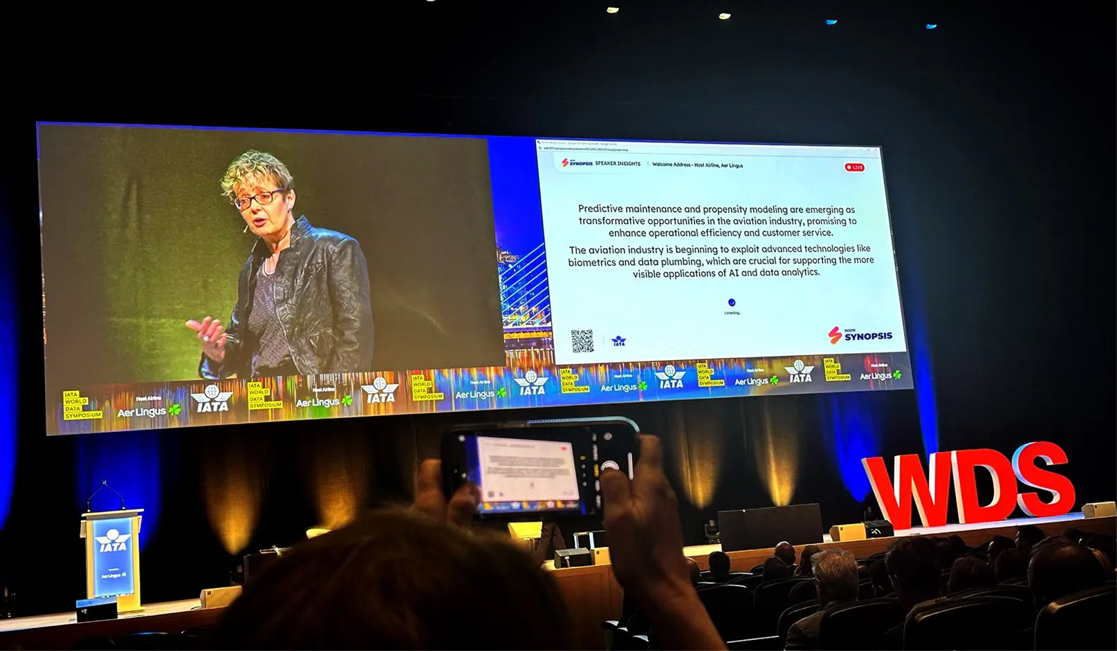 Speaker presenting on stage at IATA World Data Symposium with large screen showing presentation text on predictive maintenance in aviation and logos of Aer Lingus, IATA, and Synopsis.