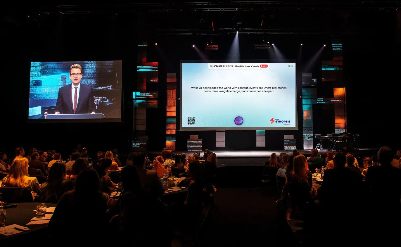 Audience seated and watching a speaker on stage in a dimly lit conference hall.