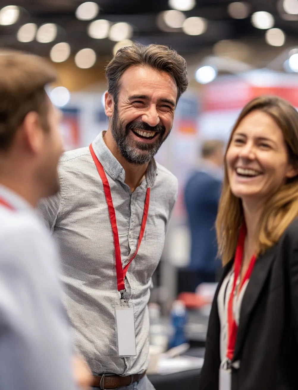 Three coworkers with red lanyards laughing and chatting in a brightly lit conference or office setting.